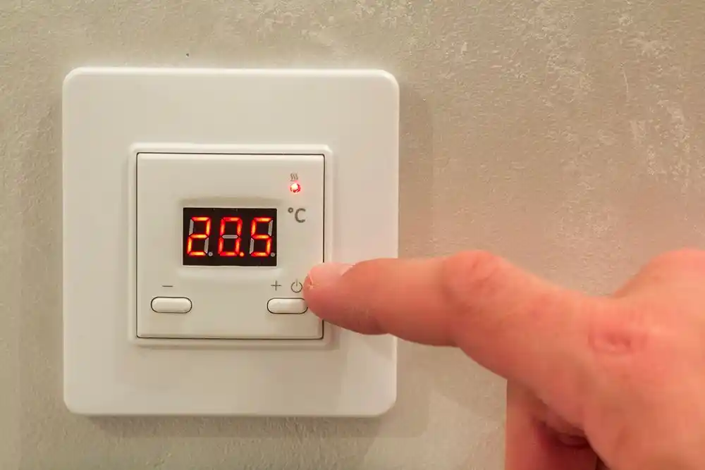 Duvara monte termostat 23,5 °C gösteriyor; bir parmak açma-kapama düğmesine basıyor
