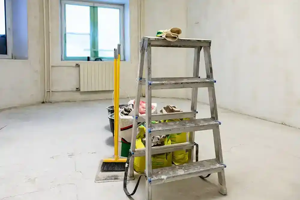 Boş bir odada, temizlik malzemelerinin yanında duran alüminyum merdiven ve sarı süpürge