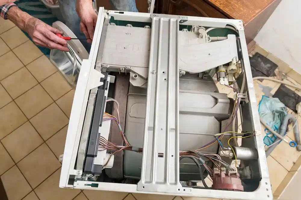 Kapağı çıkarılmış bir çamaşır makinesinin içindeki parçaları inceleyen ve tornavidayla onarım yapan kişi