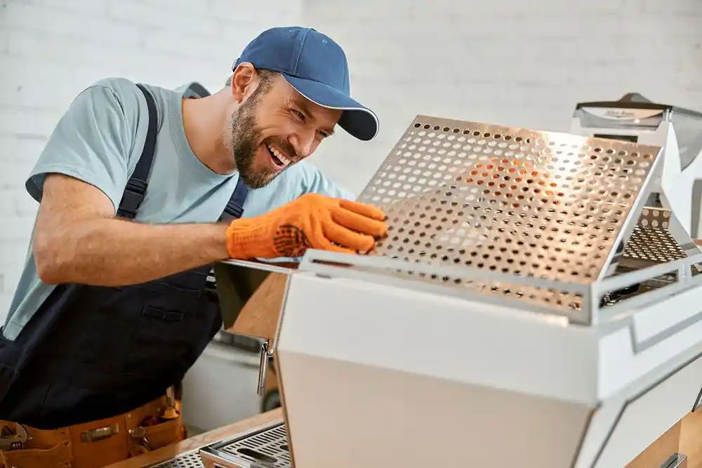 Turuncu eldiven takan bir işçi, delikli metal levhayı endüstriyel bir makineye yerleştiriyor