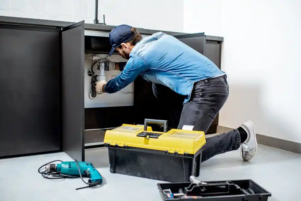 Bir teknisyen, lavabo altındaki tesisatı onarırken yanında alet çantası ve elektrikli aletler bulunuyor