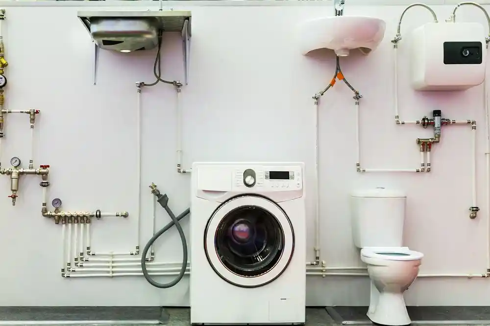 Açıkta tesisat boruları arasında çamaşır makinesi, klozet ve su ısıtıcı bulunan bir banyo tesisatı düzeni