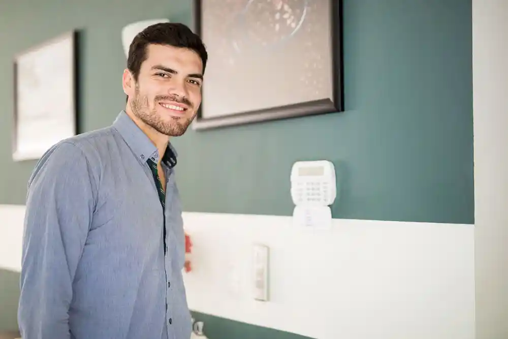Gülümseyen sakallı bir erkek, açık mavi gömlekle modern bir iç mekânda duruyor