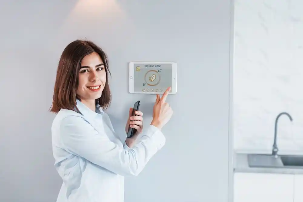 Bir kadın, elindeki telefonla duvardaki termostatı gösteriyor