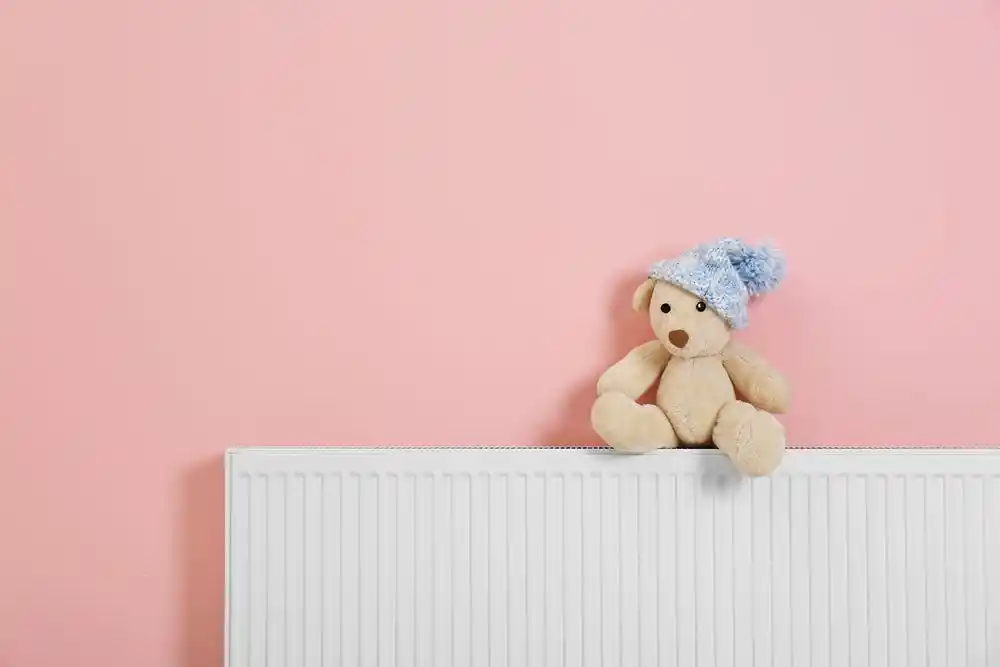 Pembe duvar önünde kalorifer peteğinin üzerinde oturan mavi bereli oyuncak ayı